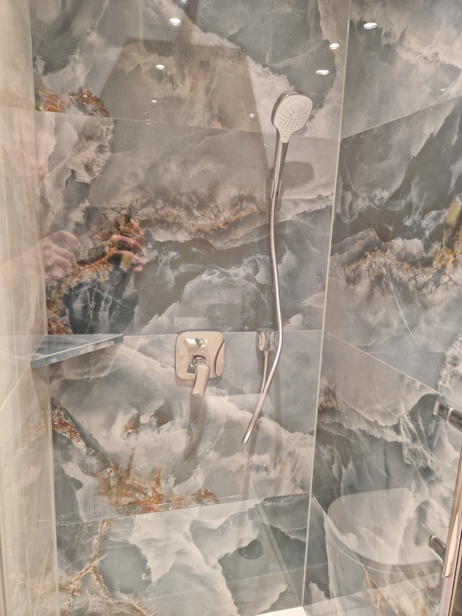 Kabina prysznicowa z armaturą i płytkami imitującymi marmur w odcieniach szarości i brązu. Widoczne odbicie osoby robiącej zdjęcie.