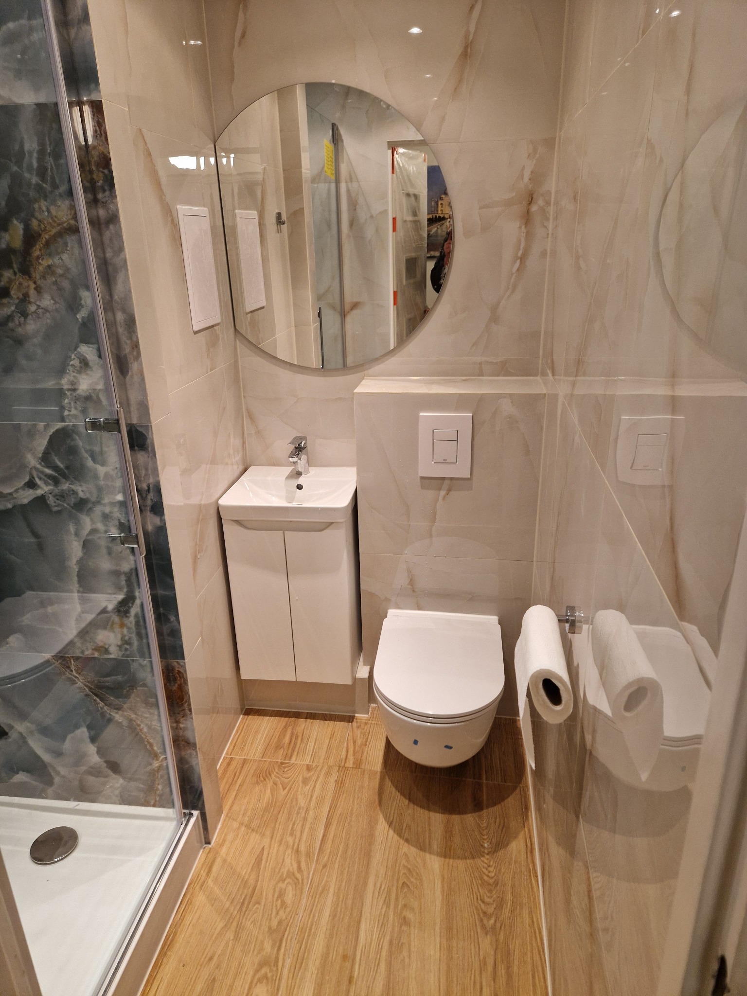 Jasna łazienka z kabiną prysznicową, umywalką z szafką, toaletą i okrągłym lustrem. Płytki imitujące marmur na ścianach i drewnopodobne na podłodze.