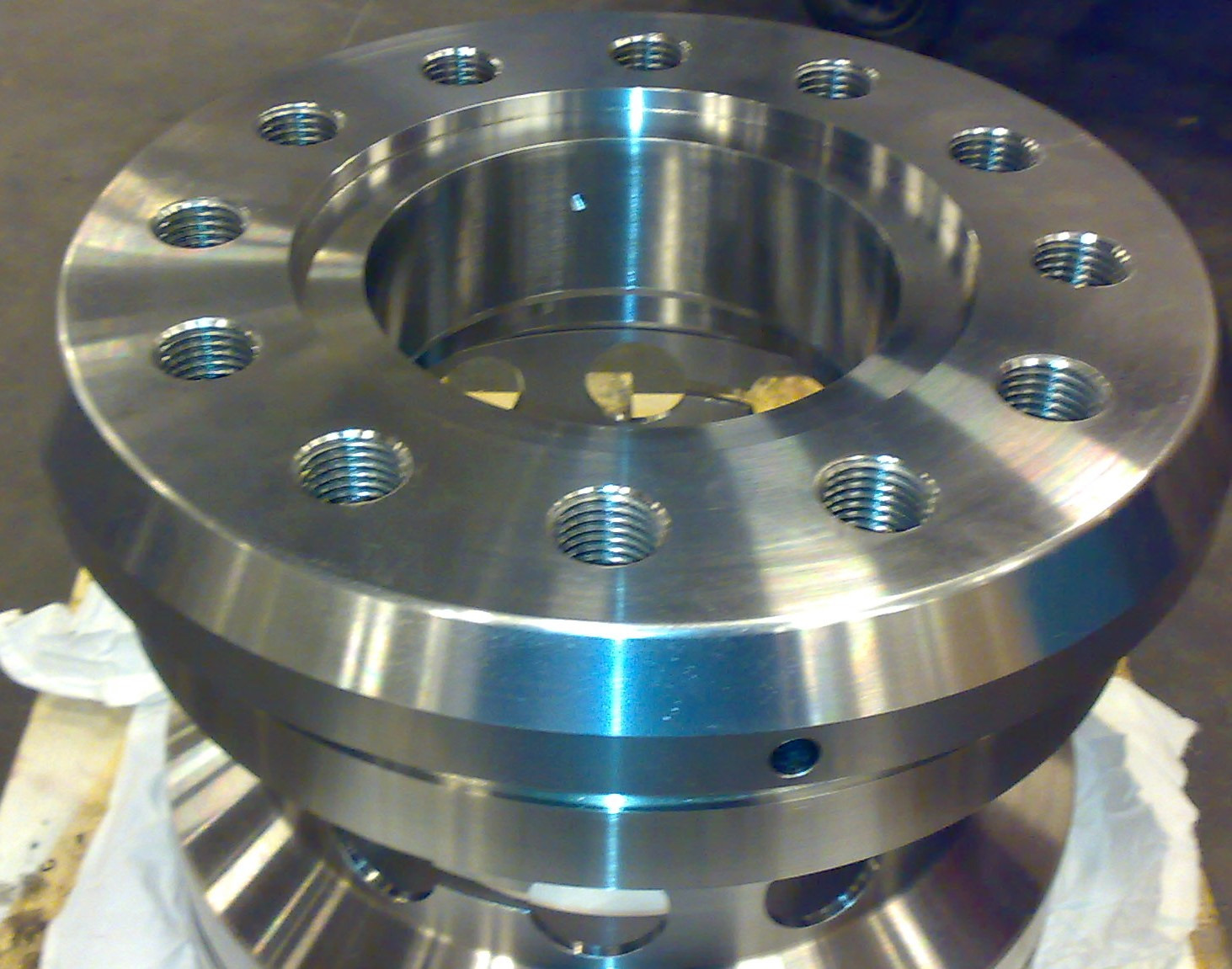 Precyzyjnie obrobiony, okrągły element metalowy z gwintowanymi otworami, widoczne refleksy światła na gładkiej powierzchni, leżący na drewnianej palecie z białym materiałem.