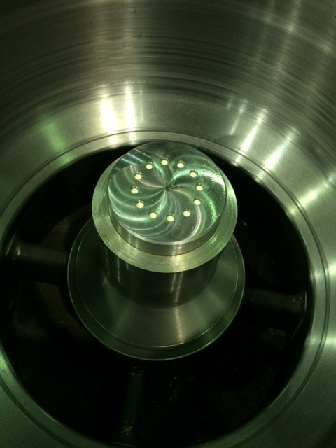 Metalowy element z otworami ułożonymi w spiralny wzór, umieszczony wewnątrz cylindrycznej metalowej obudowy, widoczne ślady obróbki.