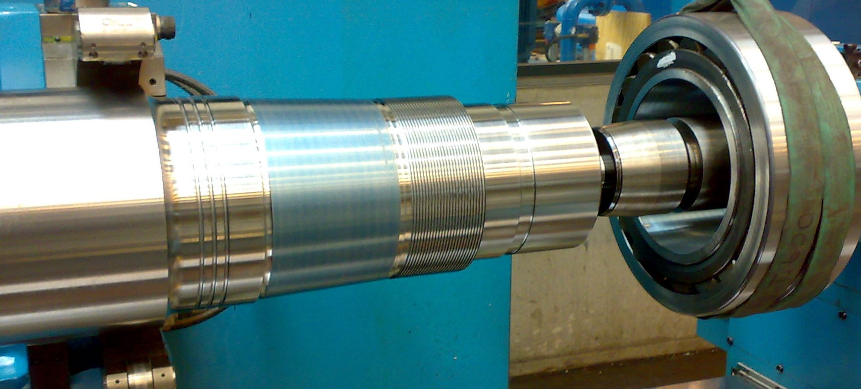 Metalowy wał z rowkami zamocowany w maszynie, prawdopodobnie podczas procesu obróbki skrawaniem lub szlifowania, z widocznym mocowaniem i elementami maszyny w tle.