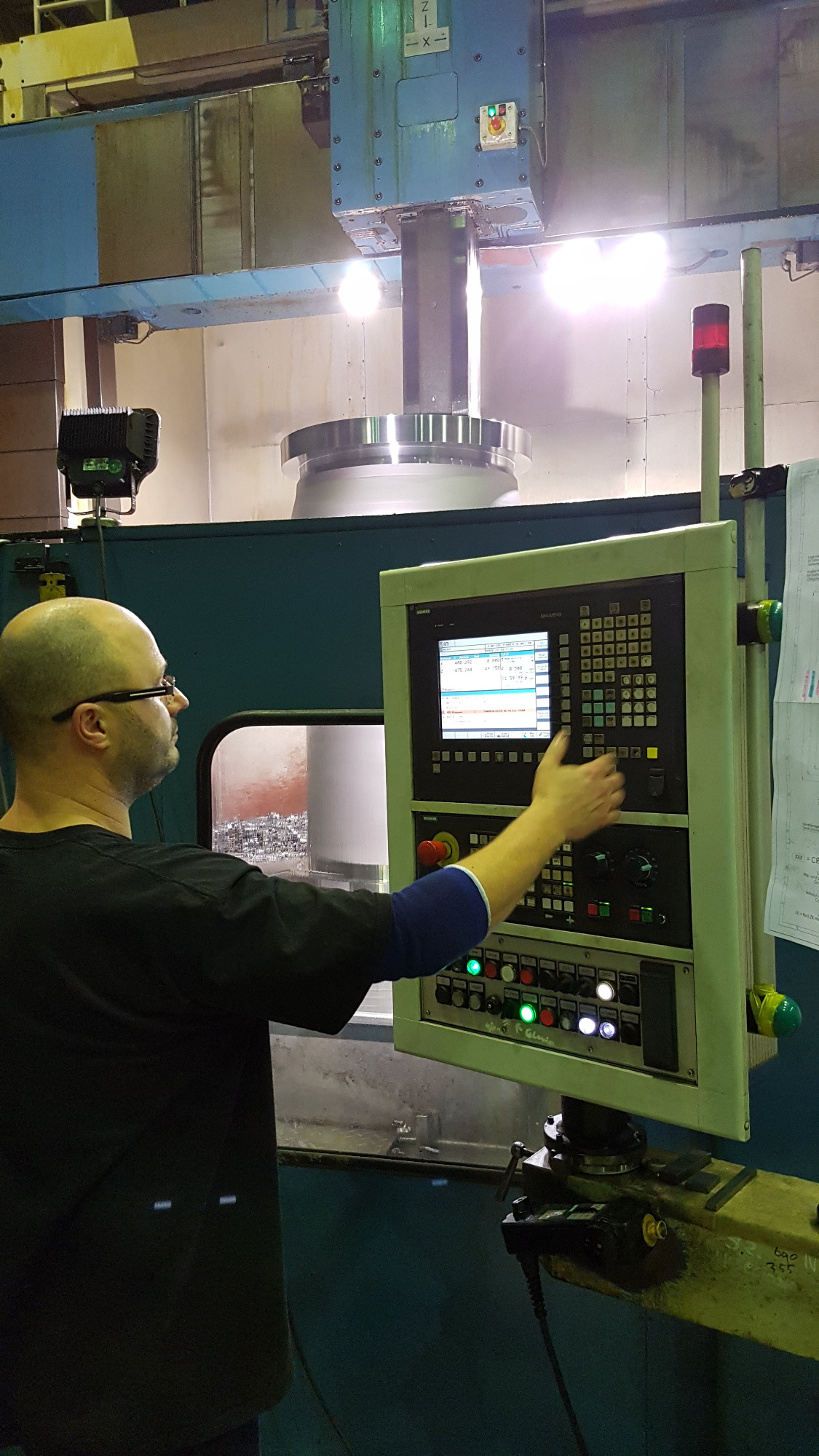 Operator wprowadza dane do panelu sterowania CNC obrabiarki podczas procesu obróbki metalu, widoczne wióry i oświetlenie przemysłowe.