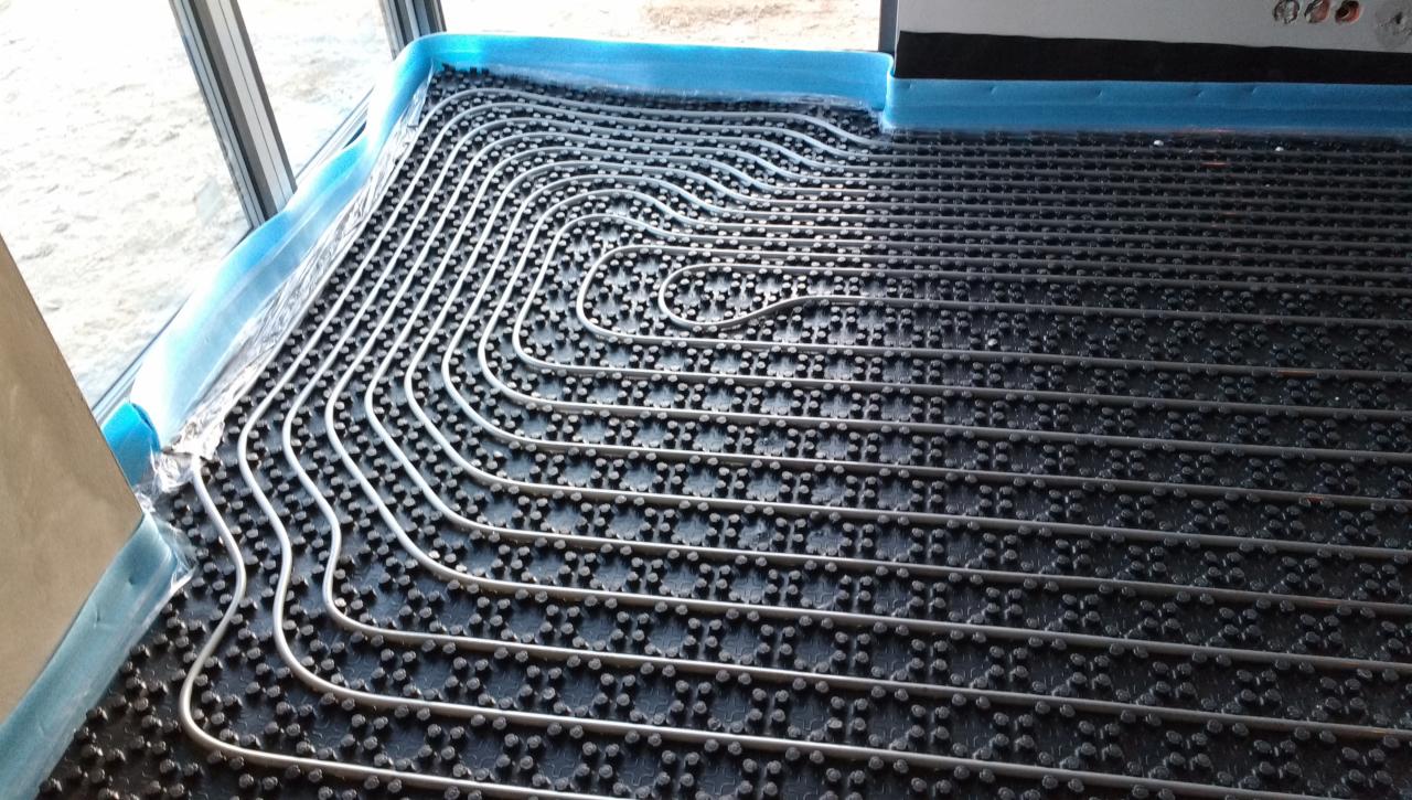 Szare rury ogrzewania podłogowego ułożone na czarnej macie z wypustkami, zabezpieczone niebieską folią przy ścianie z oknem.