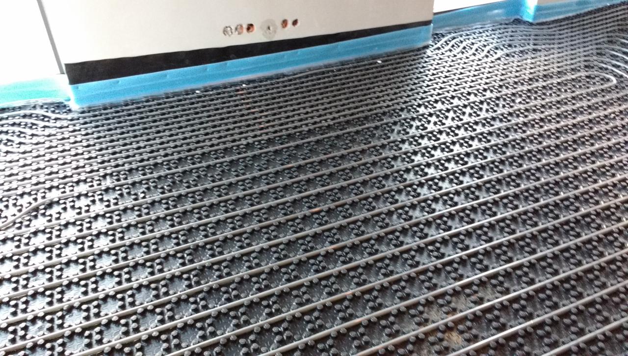 Szare rurki ogrzewania podłogowego ułożone w regularnych odstępach na czarnej macie z wypustkami, widoczne fragmenty ściany z niebieską taśmą ochronną.