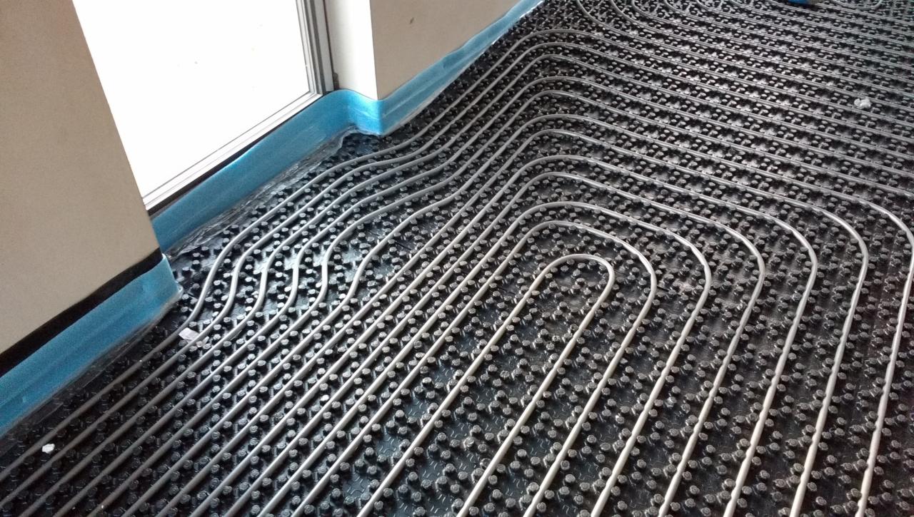 Szare rury ogrzewania podłogowego ułożone na czarnej, wypustkowej macie, zabezpieczone niebieską folią przy ścianie.