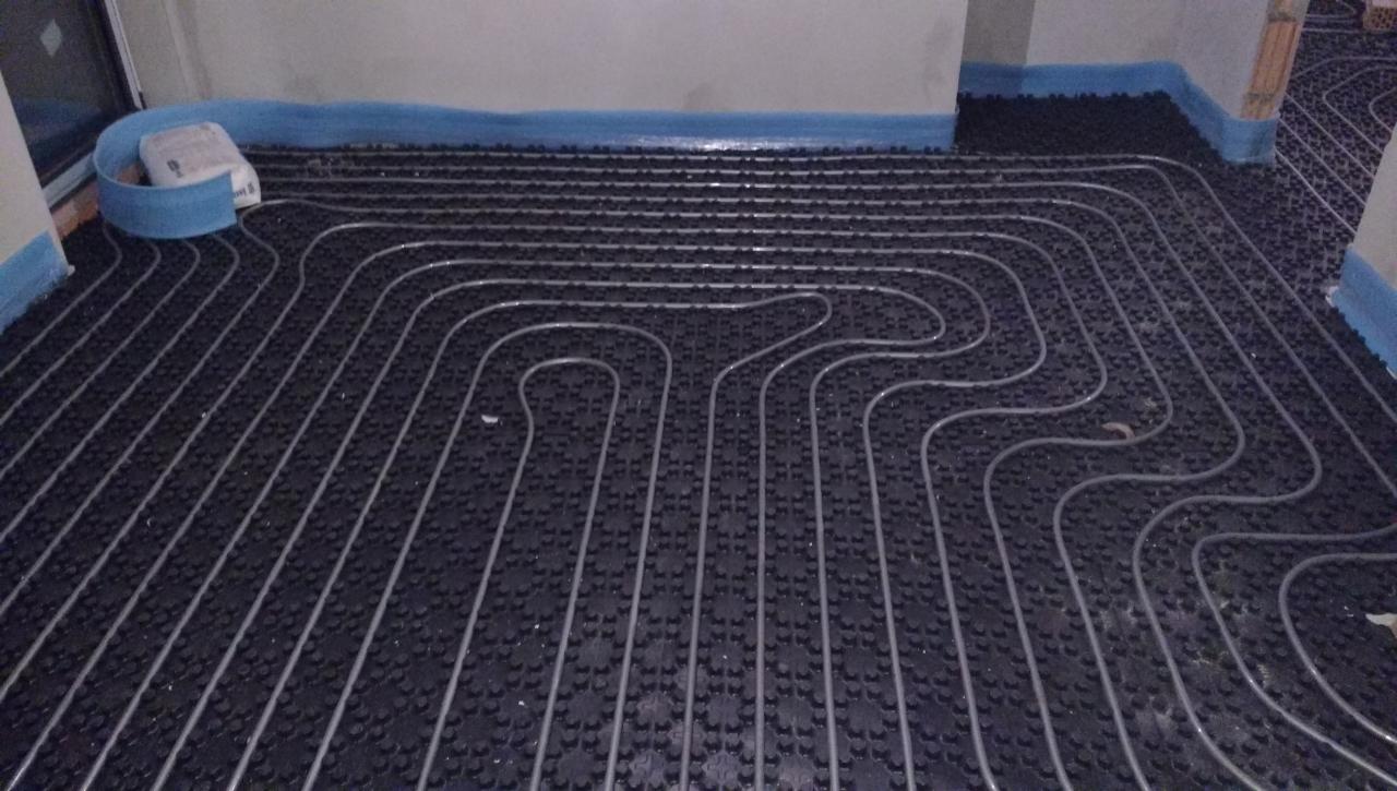 Rozłożone rury ogrzewania podłogowego na czarnej macie izolacyjnej z niebieską taśmą zabezpieczającą przy ścianach.