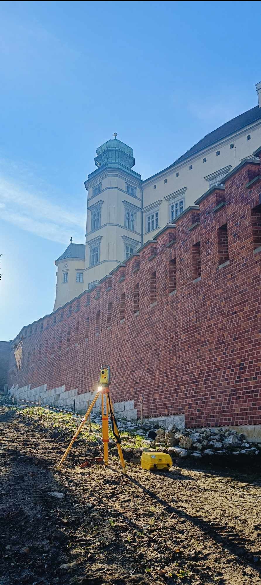 Żółty niwelator na trójnogu ustawiony na nierównym terenie z ceglanym murem i fragmentem zabytkowego budynku w tle, w słoneczny dzień.