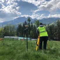 Mężczyzna w jaskrawej kamizelce, używający niwelatora na trójnogu na zielonym trawiastym terenie, w tle widoczne góry i błękitne niebo z chmurami.