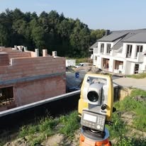 Żółty teodolit elektroniczny ustawiony na statywie na placu budowy, w tle budynek w stanie surowym otwartym oraz domy jednorodzinne w trakcie wykańczania, widoczne drzewa i błękitne niebo.