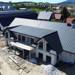 Grzędziak Budownictwo - Wznoszony dom jednorodzinny z szarego bloczka, z ukończonym dachem z blachy w kolorze grafitowym i dwoma oknami dachowymi, widok z góry na plac budowy z elementami szalunku i stertą gruzu.