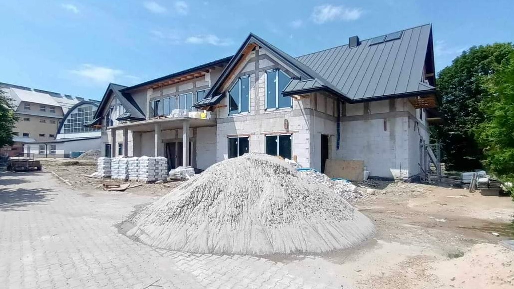 Wznoszony budynek z szarego bloczka z zamontowanymi oknami i pokrytym szarym dachem, na pierwszym planie spora sterta piasku i worki z zaprawą.