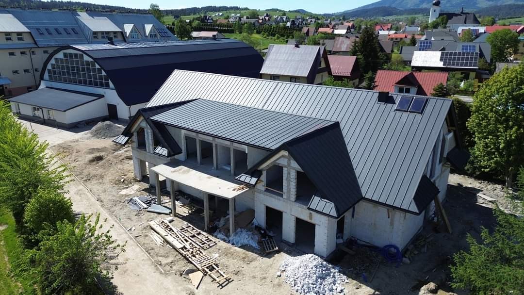 Wznoszony dom jednorodzinny z szarego bloczka, z ukończonym dachem z blachy w kolorze grafitowym i dwoma oknami dachowymi, widok z góry na plac budowy z elementami szalunku i stertą gruzu.