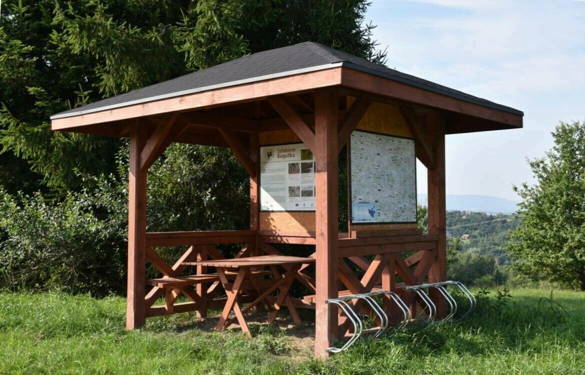 Drewniana altana z czarnym dachem na wzgórzu, wyposażona w stół z ławkami, tablice informacyjne i stojak na rowery.