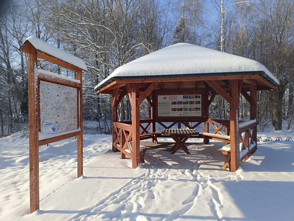 Drewniana altana z dachem pokrytym śniegiem, obok tablica informacyjna z mapą, zimowy krajobraz.