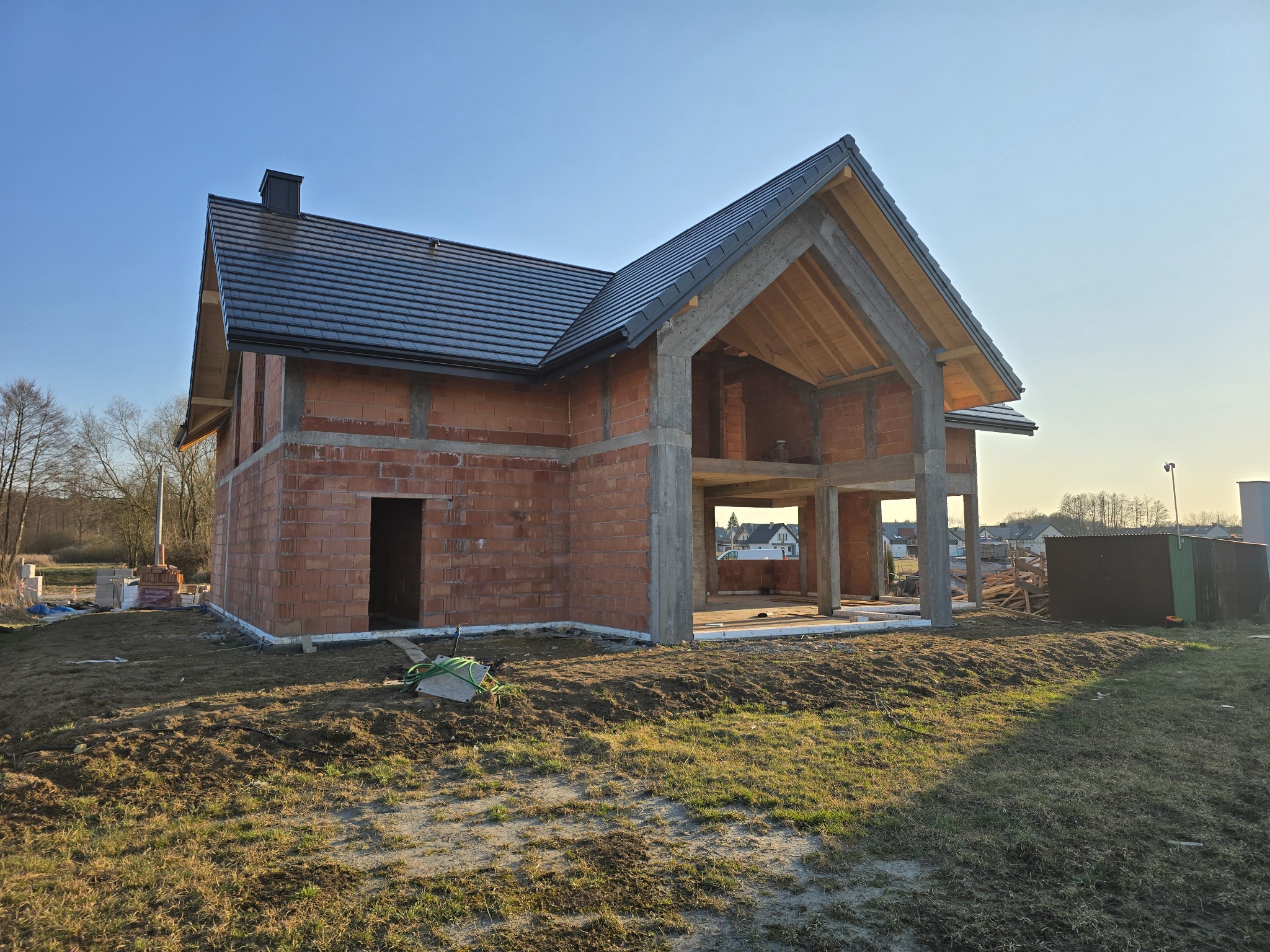 Surowy stan domu z czerwonej cegły z częściowo położonym ciemnym dachem, widoczne drewniane elementy konstrukcyjne i fundamenty, w tle zabudowa jednorodzinna.