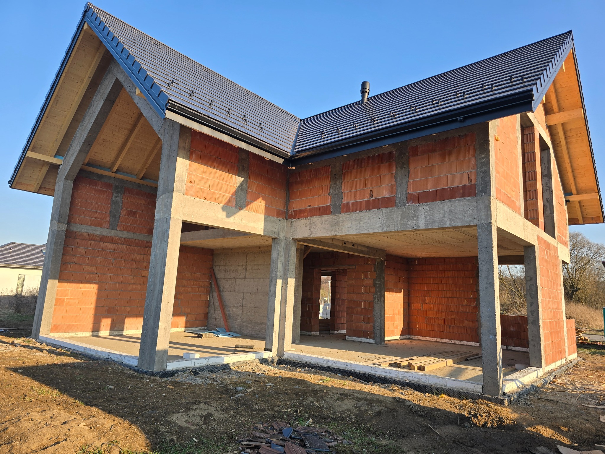 Dwukondygnacyjny budynek w stanie surowym otwartym z częściowo wykonanym ciemnym dachem, widoczne ściany z czerwonej cegły i betonowe elementy konstrukcyjne w słoneczny dzień.