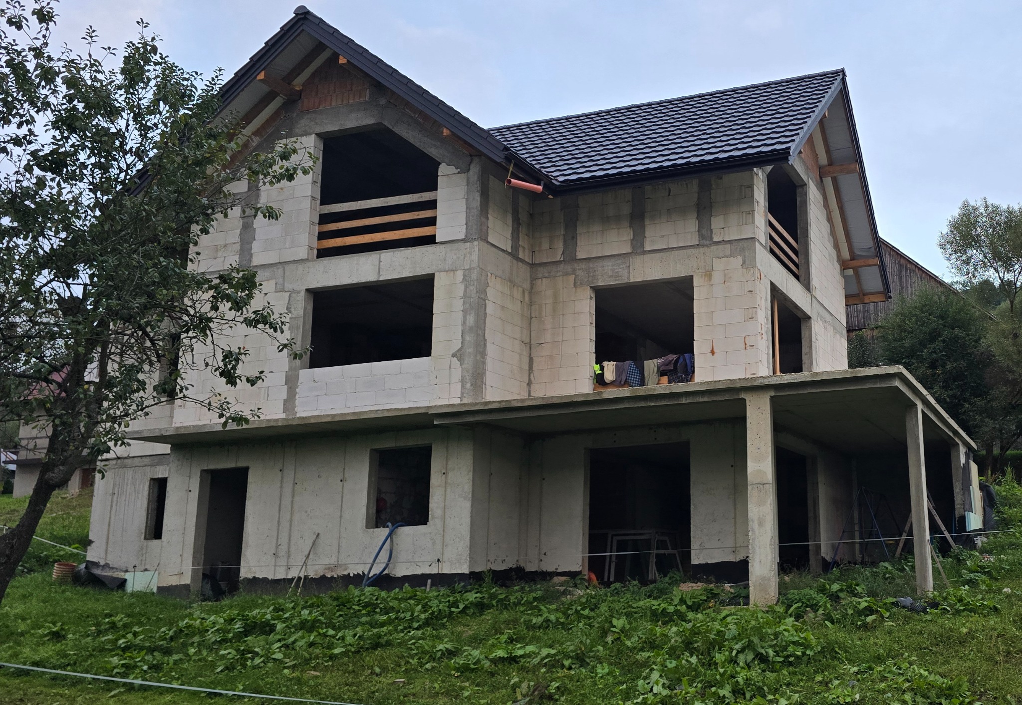 Dwukondygnacyjny dom w stanie surowym otwartym z czarnym dachem, widok z zewnątrz na tle zieleni i pochmurnego nieba. W oknach widoczne pranie.