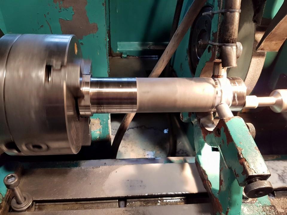Metalowy wałek obrabiany na tokarce z widocznymi opiłkami i chłodziwem, na tle zielonej, używanej maszyny.