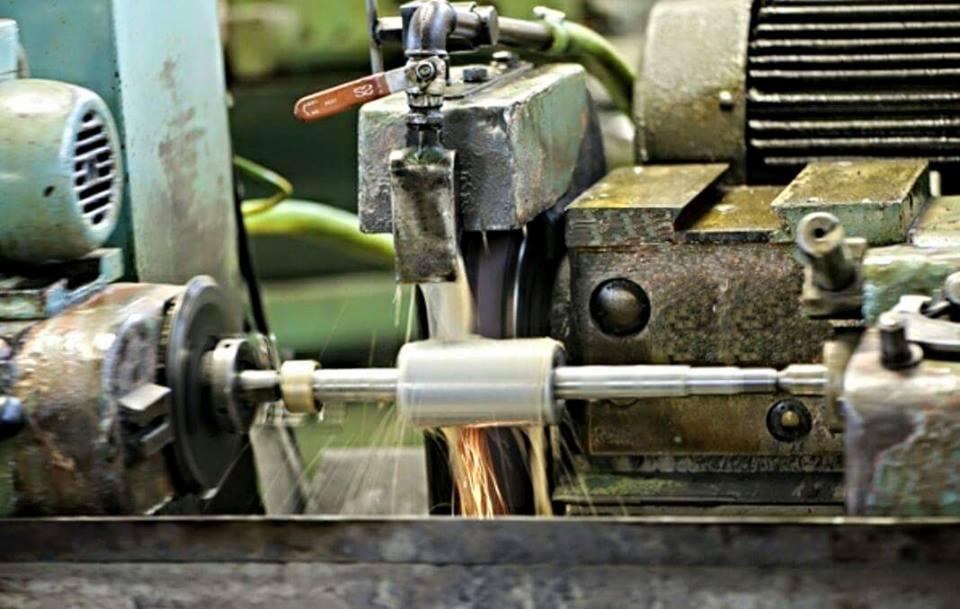 Szlifowanie metalowego wałka na szlifierce. Wióry i chłodziwo w trakcie obróbki. Widoczne elementy maszyny i obrabiany detal. Obróbka skrawaniem.