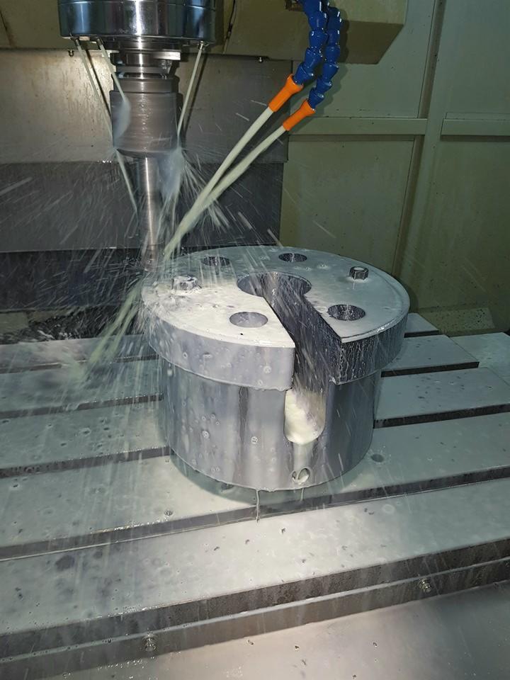 Proces obróbki metalu na maszynie CNC z chłodzeniem cieczą. Frezarka w trakcie pracy nad cylindrycznym elementem z wycięciem, widoczne wióry i płyn chłodzący.