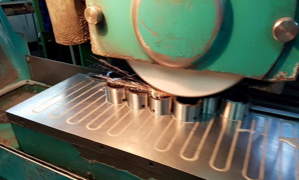 Szlifowanie metalowych tulejek na szlifierce, iskry podczas obróbki, widoczna metalowa podstawa z rowkami chłodzącymi.