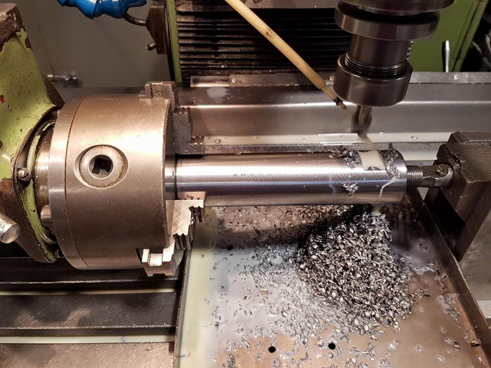 Metalowy wałek obrabiany na tokarce CNC, wióry metalowe widoczne na stole roboczym, chłodziwo spływa po obrabianym elemencie.