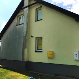 Żółta elewacja domu z widocznym fragmentem po myciu, kontrastującym z brudną częścią ściany, okna z białymi ramami, skrzynka gazowa i elektryczna na ścianie.