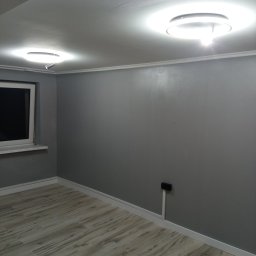 PHU Franko - Szare, świeżo pomalowane wnętrze pokoju z drewnopodobną podłogą, białymi listwami przypodłogowymi i dwoma nowoczesnymi, okrągłymi lampami sufitowymi.