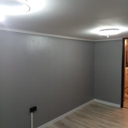 PHU Franko - Pokój po remoncie: świeżo pomalowane szare ściany, jasna podłoga imitująca drewno, białe listwy przypodłogowe i sufitowe, nowoczesne oświetlenie sufitowe.