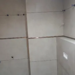 Ściana łazienki w trakcie wykańczania, z jasnymi płytkami ceramicznymi, widoczne niebieskie elementy poziomowania płytek, metalowa listwa dekoracyjna i otwór na przycisk spłuczki.