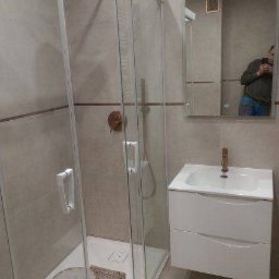 PHU Franko - Prysznic z kabiną ze szklanymi drzwiami, umywalka z szafką w łazience z beżowymi płytkami, widoczne narzędzia i wiadro z farbą sugerujące prace wykończeniowe w toku, odbicie fotografa w lustrze.