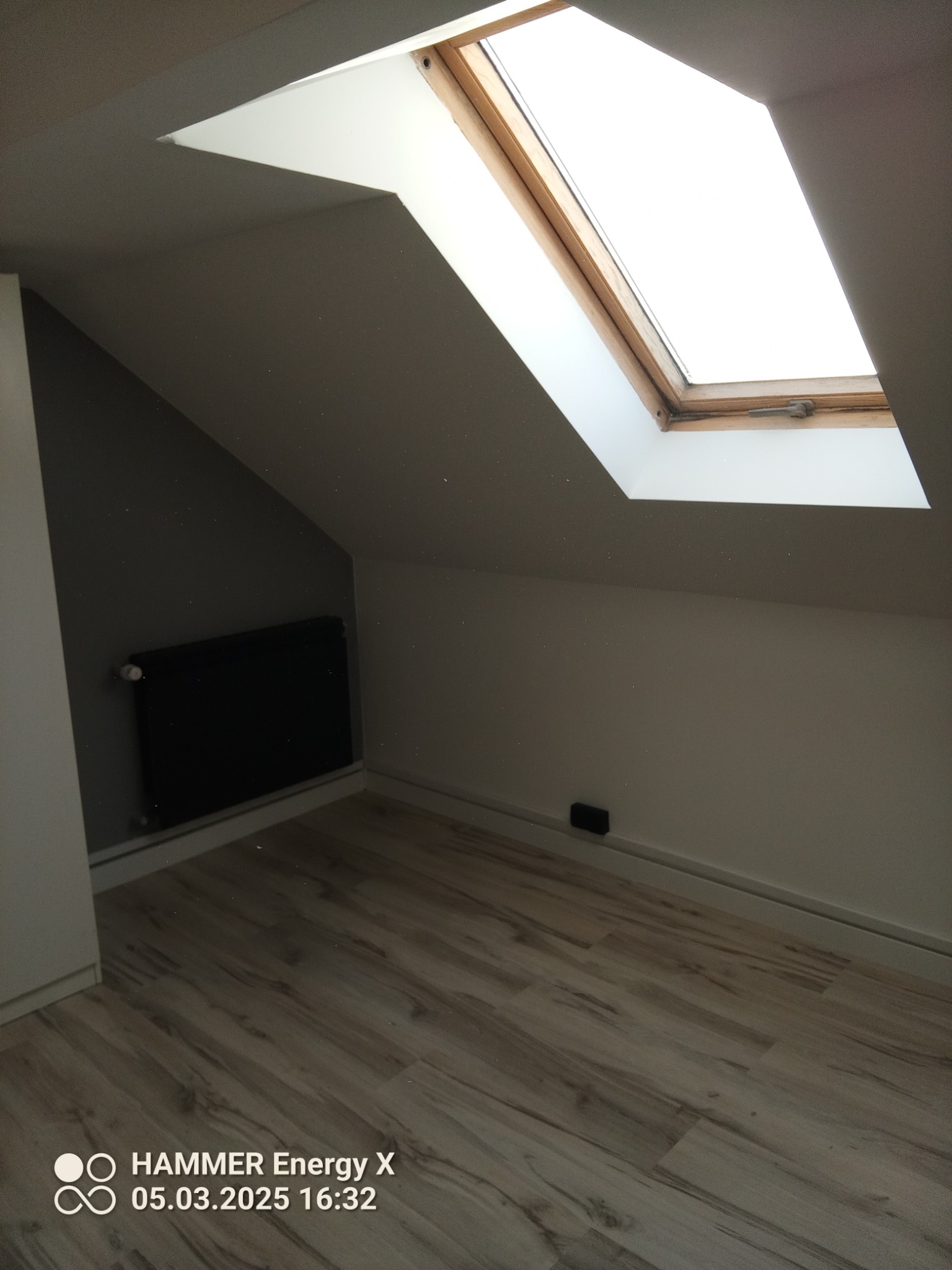 Poddasze po remoncie: wykończone ściany w odcieniach szarości i bieli, drewniana podłoga, zamontowany grzejnik i okno dachowe z drewnianą ramą.
