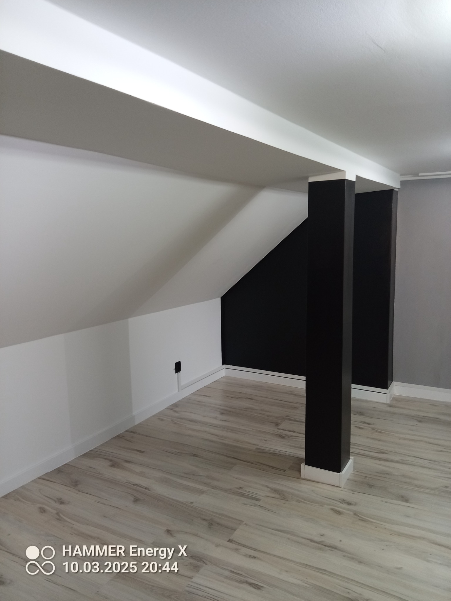 Poddasze po remoncie: białe ściany ze skosem, czarna ściana w głębi, dwie czarne kolumny z białymi bazami i kapitelami, panele podłogowe w odcieniach szarości.