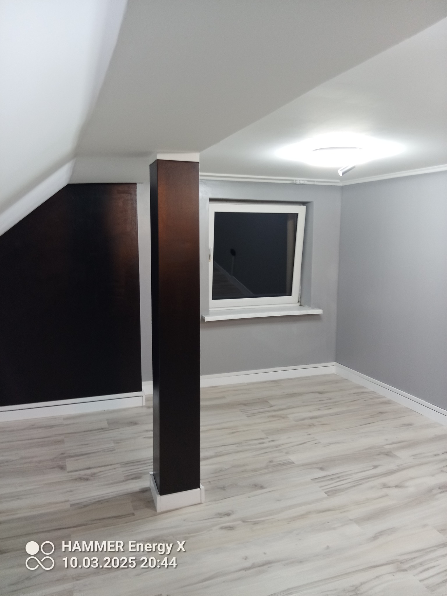 Pomalowane na szaro wnętrze poddasza z oknem, drewnianą kolumną i panelami podłogowymi imitującymi drewno.