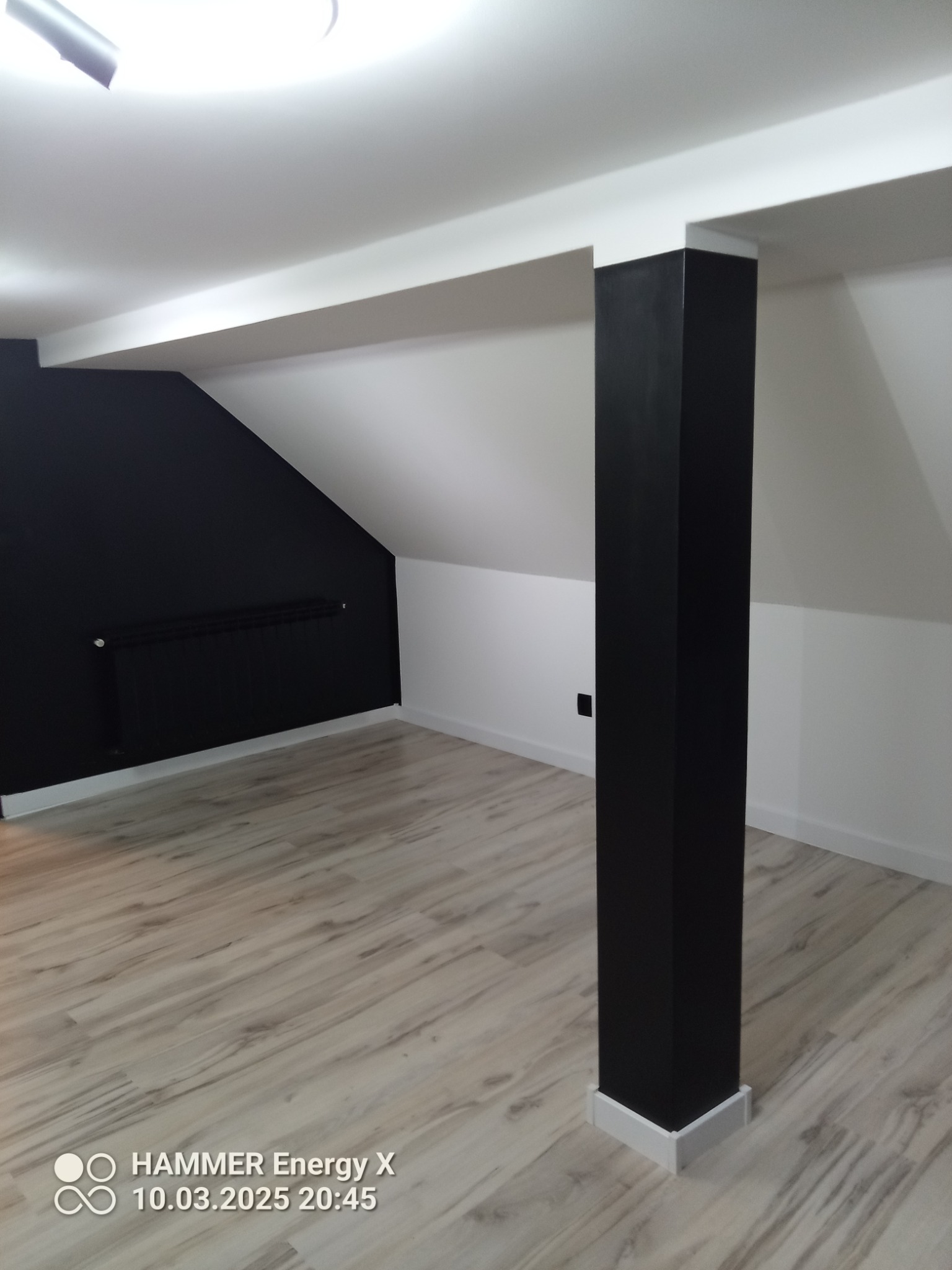 Poddasze po renowacji z czarną ścianą szczytową, grzejnikiem i drewnopodobną podłogą, wsparte czarną kolumną z białymi listwami przypodłogowymi.