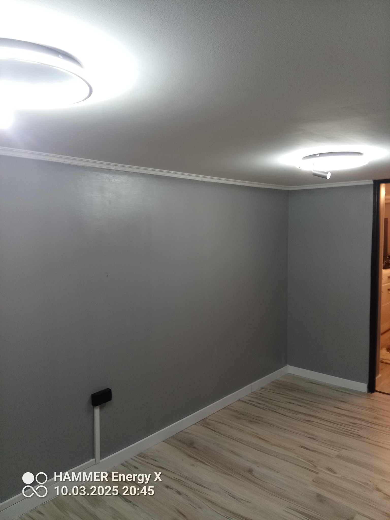 Pokój po remoncie: świeżo pomalowane szare ściany, jasna podłoga imitująca drewno, białe listwy przypodłogowe i sufitowe, nowoczesne oświetlenie sufitowe.