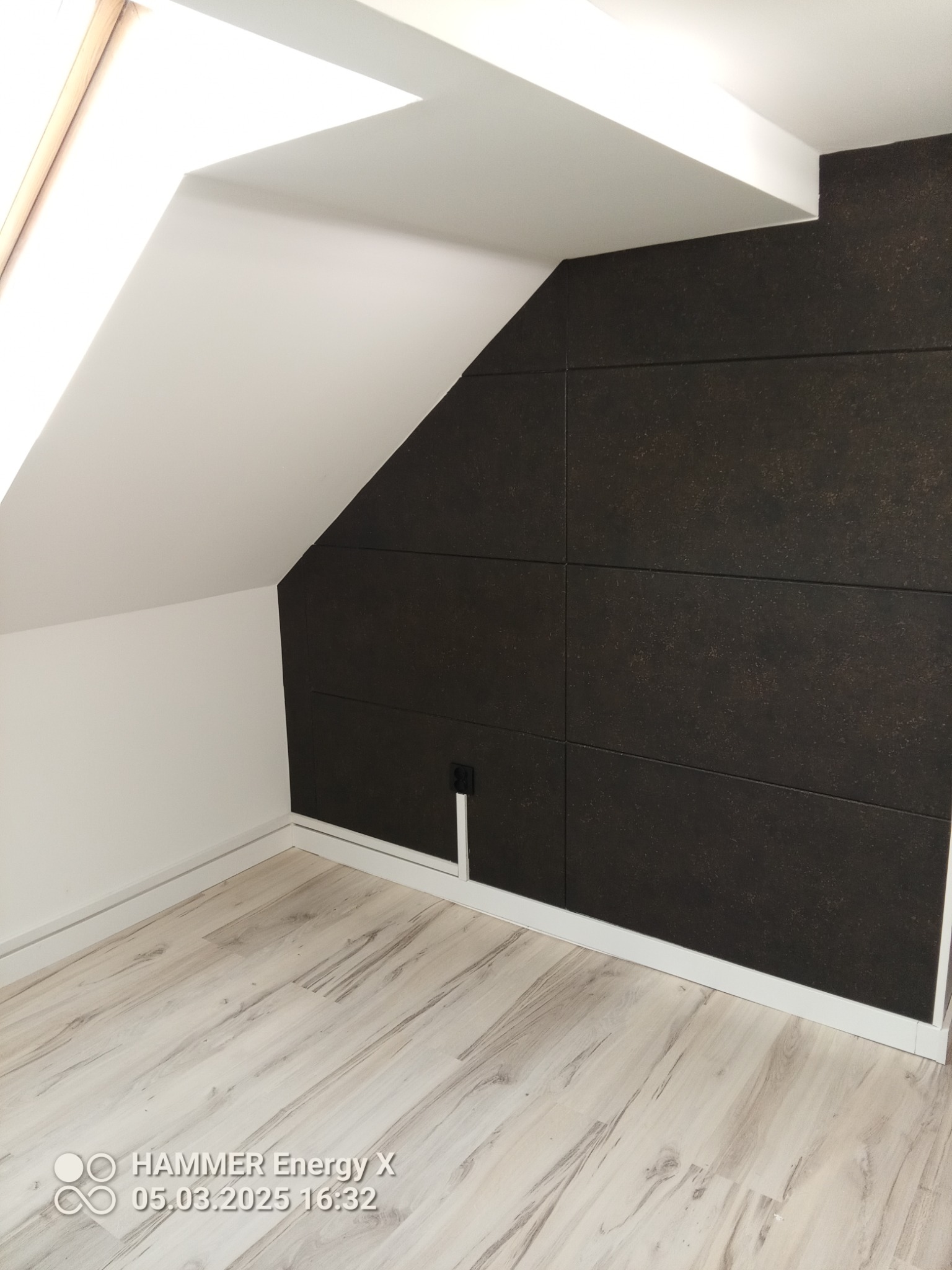 Pokój na poddaszu z panelami ściennymi w kolorze ciemnego brązu, białe ściany ze skosami, jasna podłoga i listwy przypodłogowe, widoczne gniazdko elektryczne.