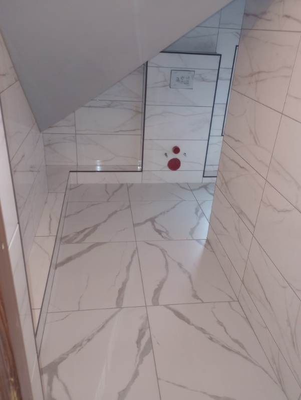 Wykończona łazienka z białymi płytkami imitującymi marmur na podłodze i ścianach, widoczne czerwone elementy instalacji wodnej i ukryty rewizyjny właz.