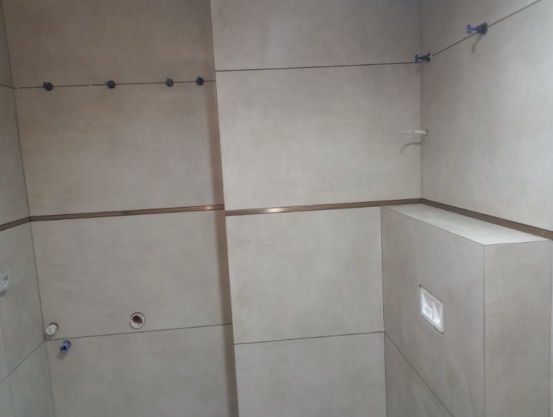 Ściana łazienki w trakcie wykańczania, z jasnymi płytkami ceramicznymi, widoczne niebieskie elementy poziomowania płytek, metalowa listwa dekoracyjna i otwór na przycisk spłuczki.