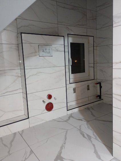 Wykończona łazienka z białymi płytkami imitującymi marmur, zabudowa geberit z przyciskiem spłukującym, okno w czarnej ramie, oraz czerwone zaślepki na rurach.