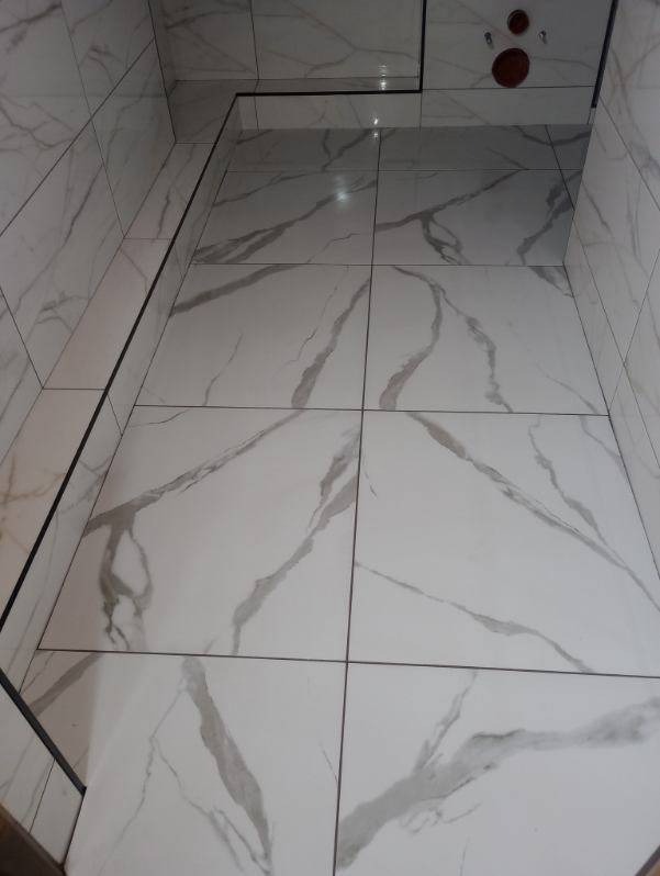 Wykończona łazienka z białymi płytkami imitującymi marmur na ścianach i podłodze, czarne fugi, widoczne przyłącza wodne.