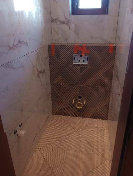 Wnętrze łazienki w trakcie remontu, widoczne płytki marmurowe na ścianach oraz dekoracyjne płytki z geometrycznym wzorem, zamontowany stelaż podtynkowy WC z pomarańczowymi taśmami montażowymi,...