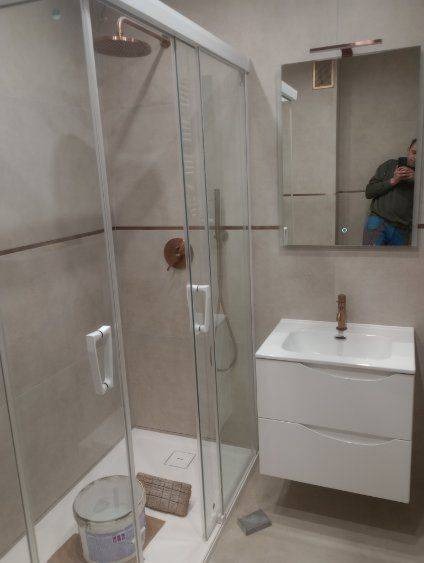 Prysznic z kabiną ze szklanymi drzwiami, umywalka z szafką w łazience z beżowymi płytkami, widoczne narzędzia i wiadro z farbą sugerujące prace wykończeniowe w toku, odbicie fotografa w lustrze.
