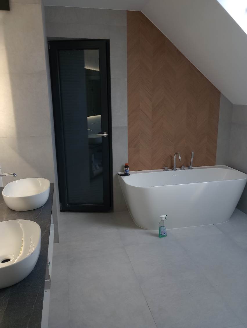 Nowoczesna łazienka na poddaszu z wolnostojącą białą wanną, dwoma umywalkami nablatowymi na ciemnym blacie i ścianą wykończoną drewnopodobną mozaiką w jodełkę.