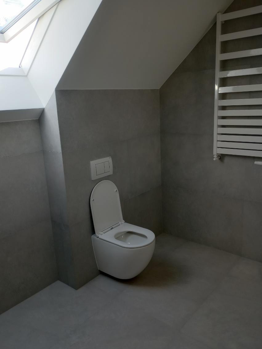 Nowoczesna toaleta podwieszana w szarej łazience na poddaszu, widoczny grzejnik drabinkowy i okno dachowe.