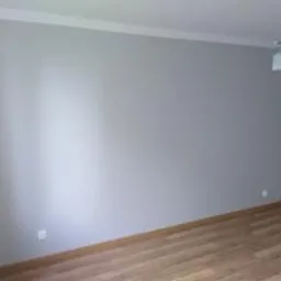 Pomalowane na szaro wnętrze pokoju z drewnianą podłogą, białą listwą przypodłogową i prostym oświetleniem sufitowym.