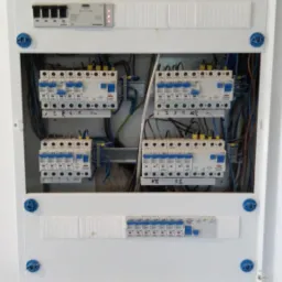 Otwarta, biała skrzynka rozdzielcza z zainstalowanymi bezpiecznikami i przewodami. Widoczne naklejki z oznaczeniami na poszczególnych modułach.