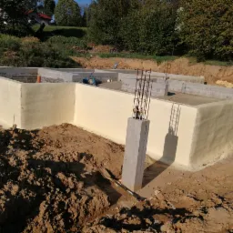 Fundament budynku po ociepleniu pianką PUR, widoczne zbrojenia i ziemia, w tle dom i drzewa.