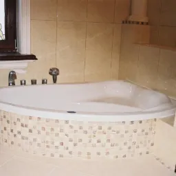 Wykończona, narożna wanna z mozaiką w beżach i brązach, armatura chromowana, jasne płytki na ścianach i podłodze. Okno z witrażem w tle.