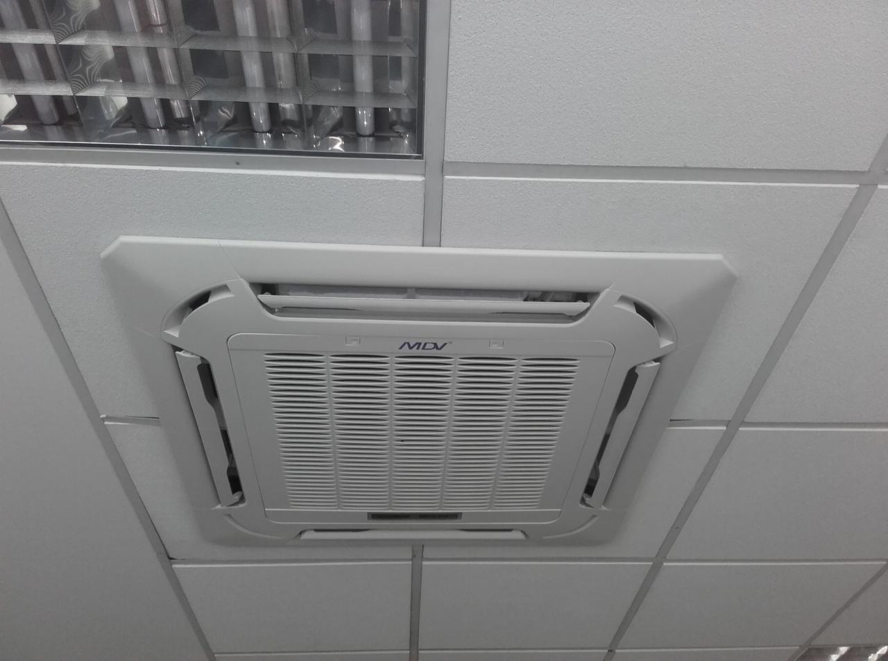 Kasetonowa jednostka klimatyzacji MDV, zamontowana w suficie podwieszanym z widocznymi elementami oświetlenia. Biała kratka nawiewna i obudowa.